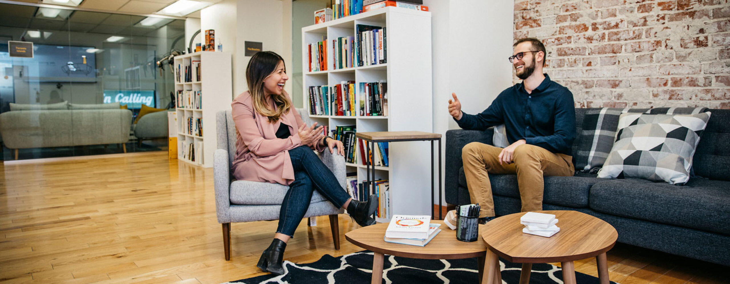 Two colleagues are having a conversation in a modern office lounge with bookshelves and cozy seating.