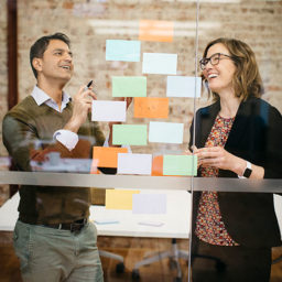 Two colleagues brainstorming with sticky notes on a glass wall.