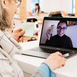 A woman in a virtual meeting, holding a sticky note while a colleague on screen engages.