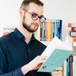 A bearded man wearing glasses and a navy blue shirt is reading a book in front of a colorful bookshelf.