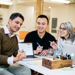 Three colleagues are collaborating at a table, discussing notes and using a laptop in a modern office space.