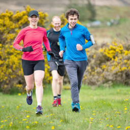 A group of three people jogging outdoors in a grassy field with yellow flowers.