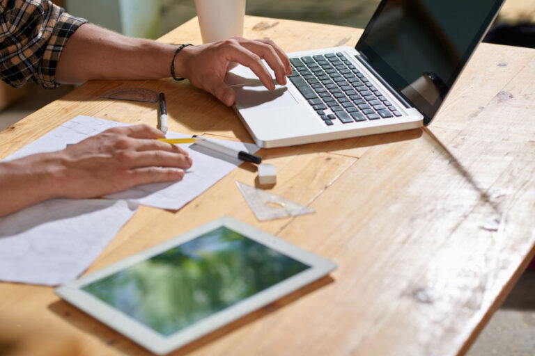 A person is working on designing a learning experience with a laptop, tablet, and sketches on a wooden desk.